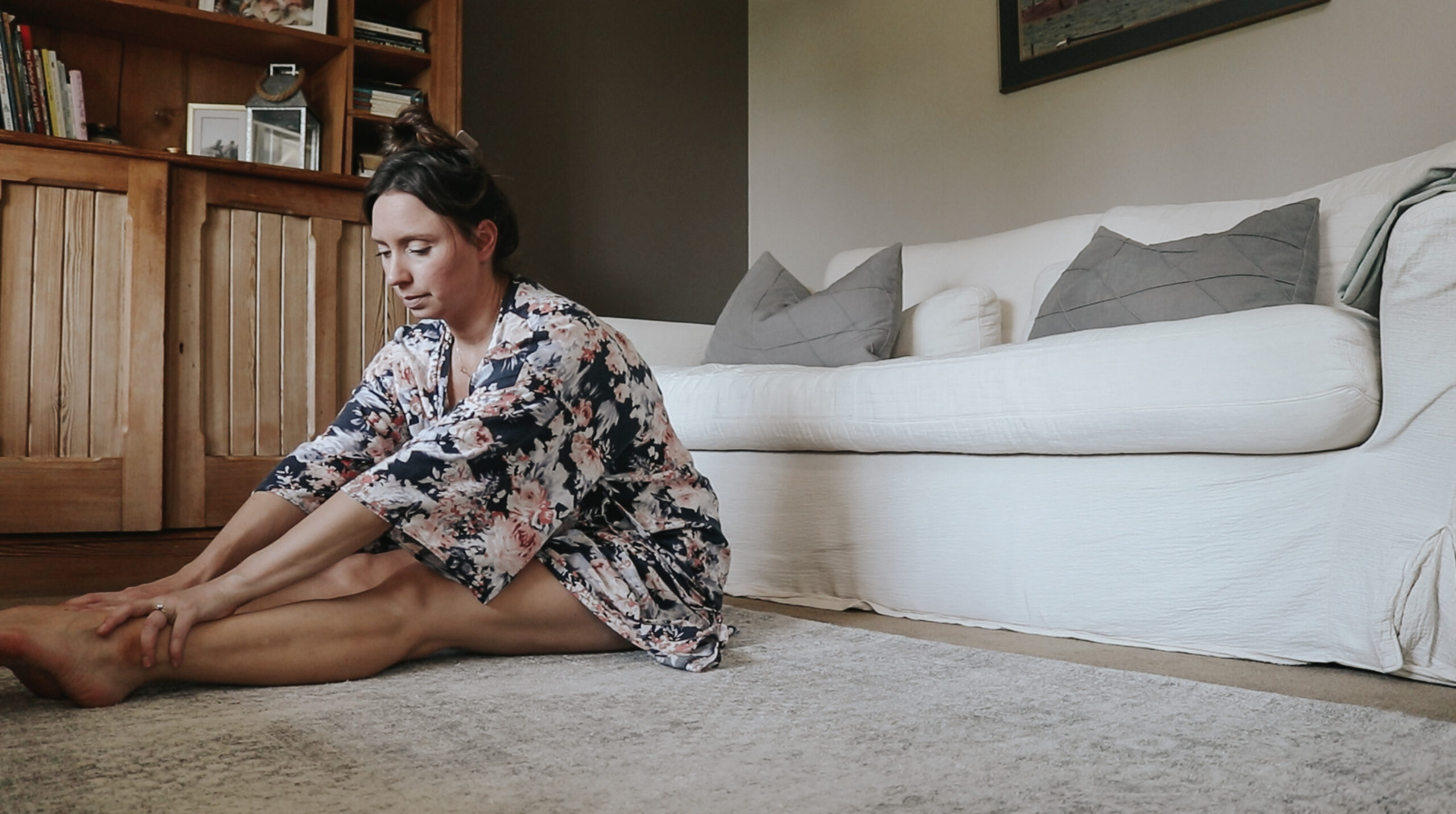 Christian homemaker sits on her living room floor wearing a floral robe and stretching forward with her arms extended and her legs together.
